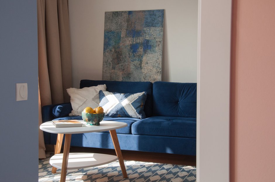 В гостиной стены в основном белые, и акцент сделали на ярко-синем диване. Его дополняют ковер, текстиль и декор также в оттенках синего и голубого.
Особое внимание стоит обратить на картину художника Ольги Шагиной, которая дополнила интерьер.